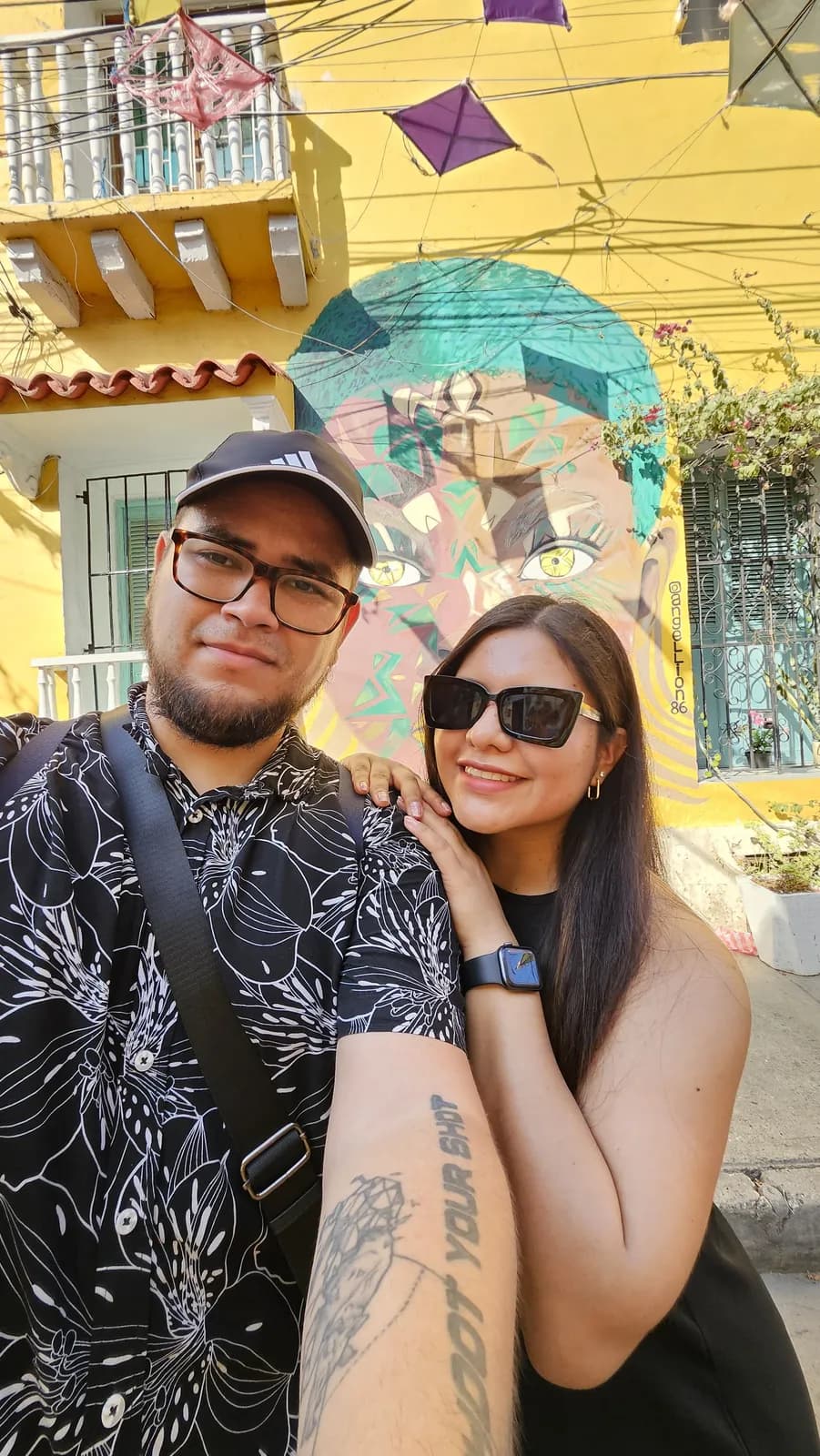 Ariadna y Rogelio en una selfie frente a un mural colorido en una calle de casas amarillas